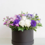 A flower arrangement with purple , white roses with chrysanthemum in a black round box