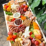 This platter serves up to 6 people with a variety of flavors. It includes five cheeses: smoked scamorza, cheddar, herb & garlic neufchatel, chili pepper neufchatel, and pistachio-cranberry cheese ball. Paired with cold cuts like beef mortadella, chicken ham, smoked beef, and chicken mortadella, it also features fresh fruit, nuts, olives, crackers, hummus dip, chutney, and a dried orange and rosemary garnish for a perfect balance of savory, sweet, and tangy.