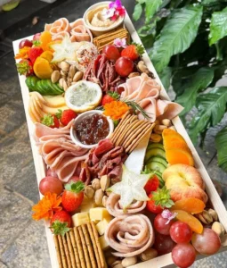 This platter serves up to 6 people with a variety of flavors. It includes five cheeses: Smoked Scamorza, Cheddar, Herb & Garlic Neufchatel, Chili Pepper Neufchatel, and Pistachio-Cranberry Cheese Ball. Paired with cold cuts like Beef Mortadella, Chicken Ham, Smoked Beef, and Chicken Mortadella, it also features fresh fruit, nuts, olives, crackers, hummus dip, chutney, and a dried orange and rosemary garnish for a perfect balance of savory, sweet, and tangy.